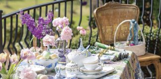 An outdoor table dressed in a floral, multicolored tablecloth is adorned with pink and purple blooms and porcelain china charms, all perfect for celebrating Easter.
