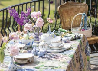 An outdoor table dressed in a floral, multicolored tablecloth is adorned with pink and purple blooms and porcelain china charms, all perfect for celebrating Easter.