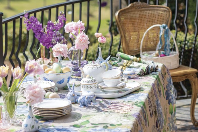 An outdoor table dressed in a floral, multicolored tablecloth is adorned with pink and purple blooms and porcelain china charms, all perfect for celebrating Easter.
