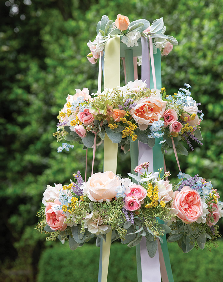 A fully dressed maypole draped in pastel ribbon, bedecked with yellow, violet, and pink flowers and emerald-green leaves.