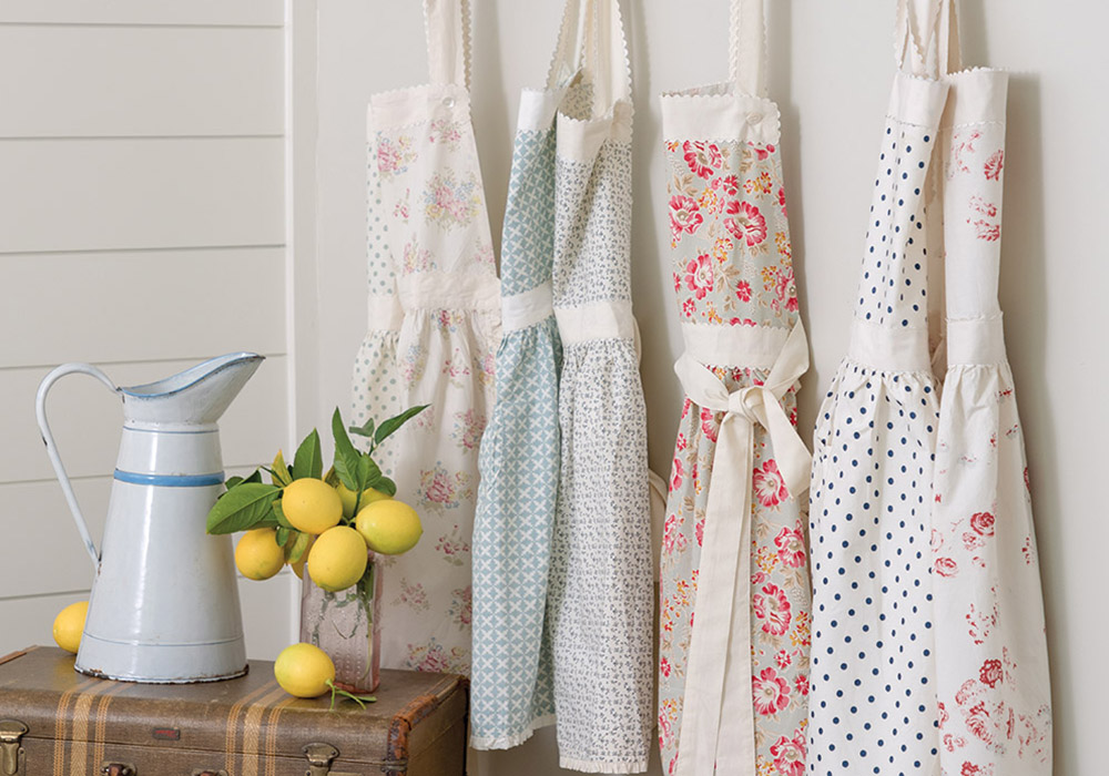 Homespun linen kitchen aprons hang against a white wood panel wall. A water pitcher and lemons nestle on a suitcase nearby.