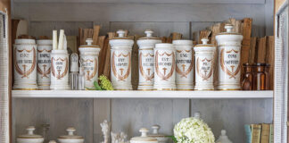 A collection of French apothecary jars is displayed on a shelf among sweet flowers and seashells.