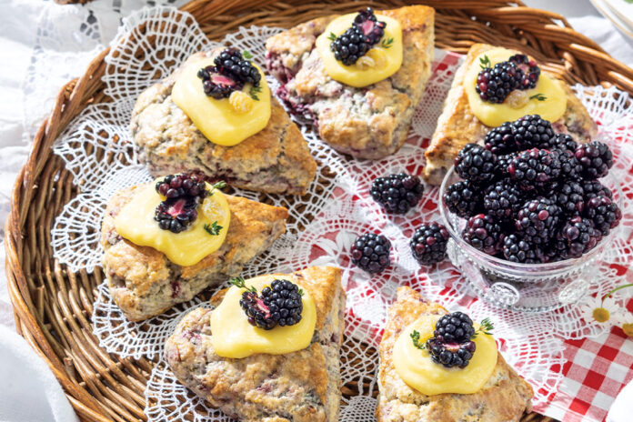 Blackberry-Thyme Scones nestle around a bowl of fresh blackberries. The pastries rest on a whicker tray, dappled in lace and red gingham.