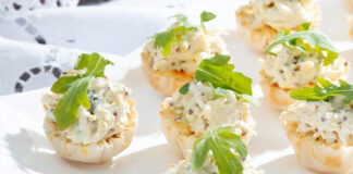 Mustard Chicken Salad Cups, accessorized in green arugula leaves, huddle on white plate, resting on a sunny painted white tablecloth.