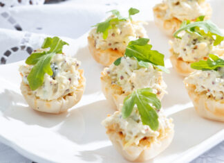 Mustard Chicken Salad Cups, accessorized in green arugula leaves, huddle on white plate, resting on a sunny painted white tablecloth.