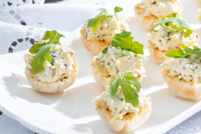Mustard Chicken Salad Cups, accessorized in green arugula leaves, huddle on white plate, resting on a sunny painted white tablecloth.