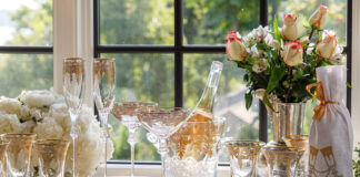 Arrangement of Arte Italica Vetro Gold glassware and white and pink flowers with a bucket of champagne and a wine bottle wrapped in a Crown Linen Designs embroidered wine bag.