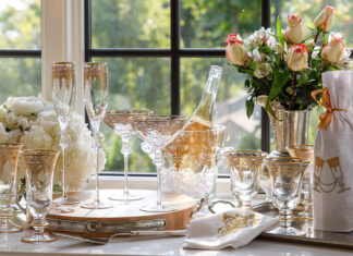 Arrangement of Arte Italica Vetro Gold glassware and white and pink flowers with a bucket of champagne and a wine bottle wrapped in a Crown Linen Designs embroidered wine bag.