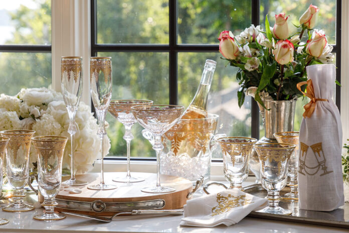 Arrangement of Arte Italica Vetro Gold glassware and white and pink flowers with a bucket of champagne and a wine bottle wrapped in a Crown Linen Designs embroidered wine bag.