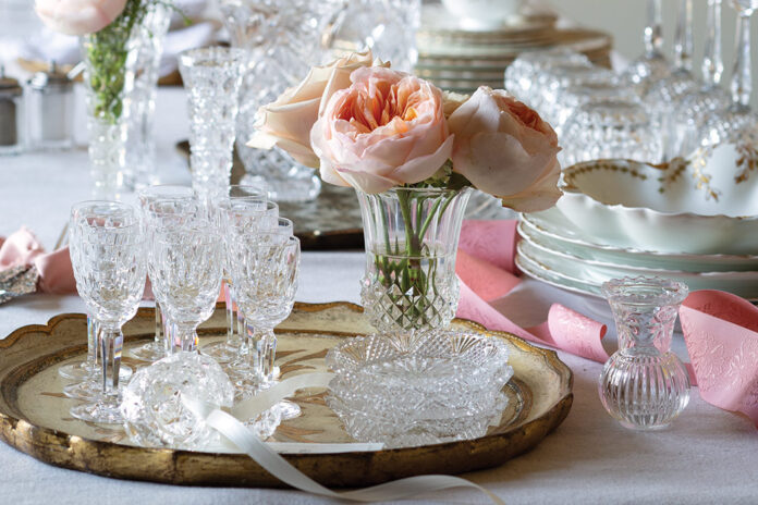 A collection of crystalware is placed atop the table for cleaning, but the work is made into a lovely display with sprays of fresh pink peonies and curls of ribbon.