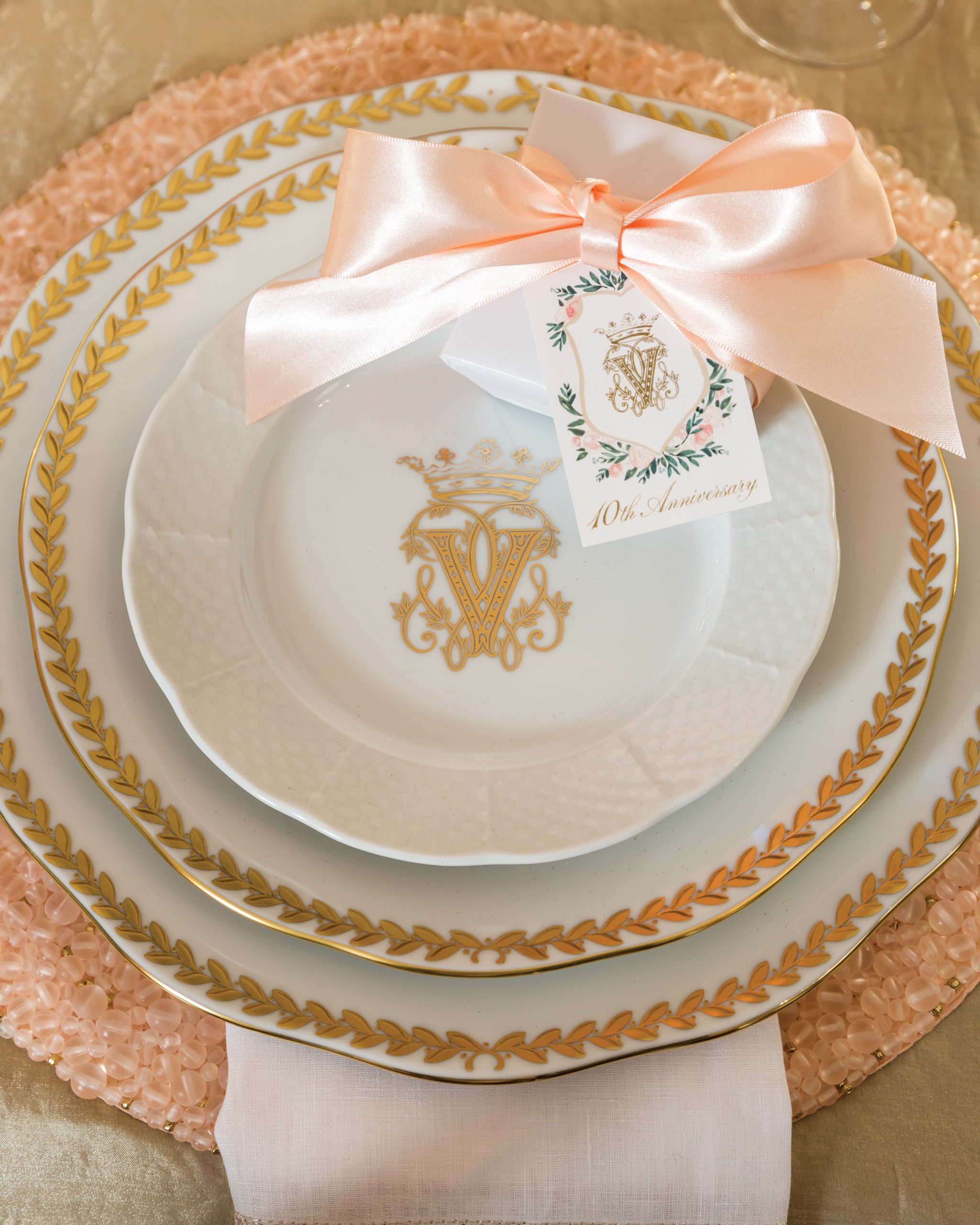 Pink and gold trimmed tiered china plates on a woven pink table mat, topped with a pink silken bow and lavender embossed seating card with 10th Anniversary across the bottom.