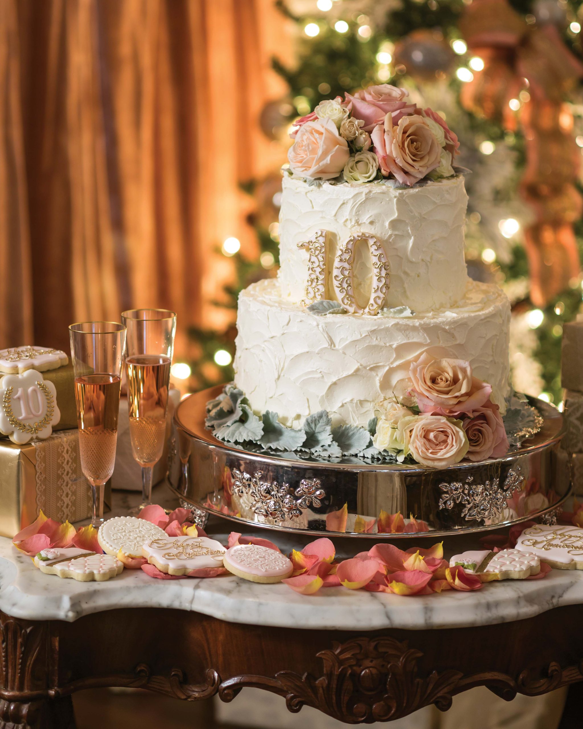 A tiered cake crowned with roses, decked with a celebratory "10" on the second tier, then lined in roses and icing on the third. Rose petals, anniversary-themed cookies, and bubbly rose in two champagne flutes gather to celebrate.
