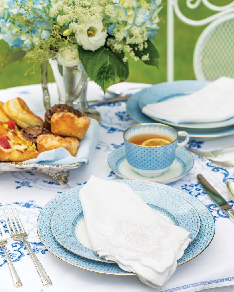 Blue and white china alights upon a graceful outdoor table, with a breadbasket of French pastries awaiting in the background.