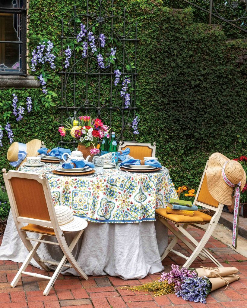 In a sunny ivy-clad courtyard inspired by Italy, a sunny table is laden with brightly patterned linens, vibrant flowers, and sun hats to shade guests’ faces.