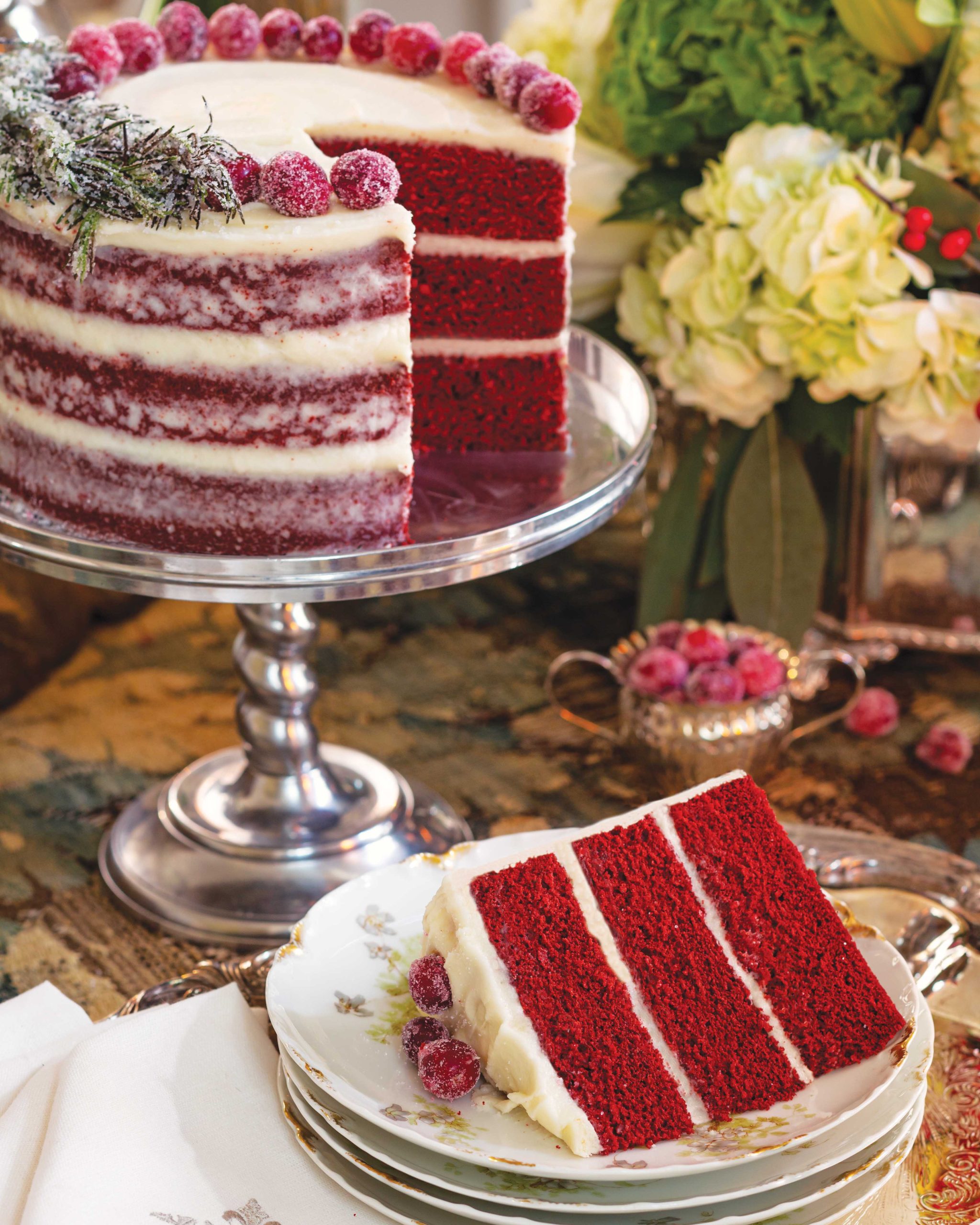 Red velvet cake topped with sugared cranberries awaits devouring, with a simple slice cut and plated.