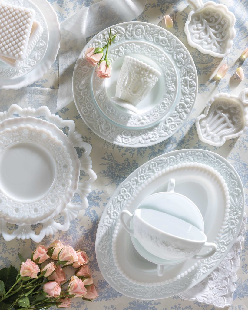 Viewed from above, a mingling of various patterns of vintage milk glass rests upon the table.