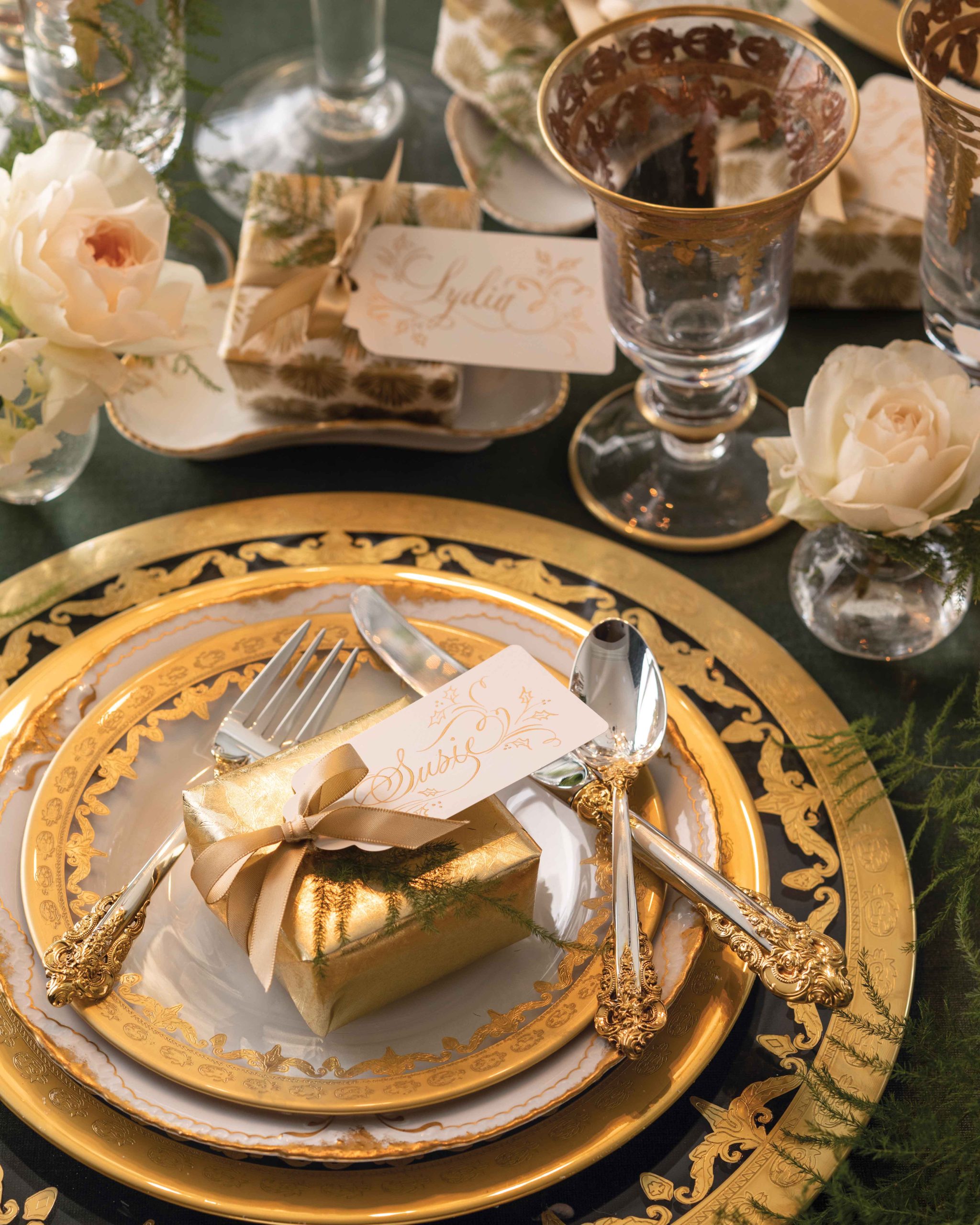 Exquisitely ornate gold plates, silverware, and goblets sit atop this table alongside small white rosebud and little wrapped Christmas gifts.