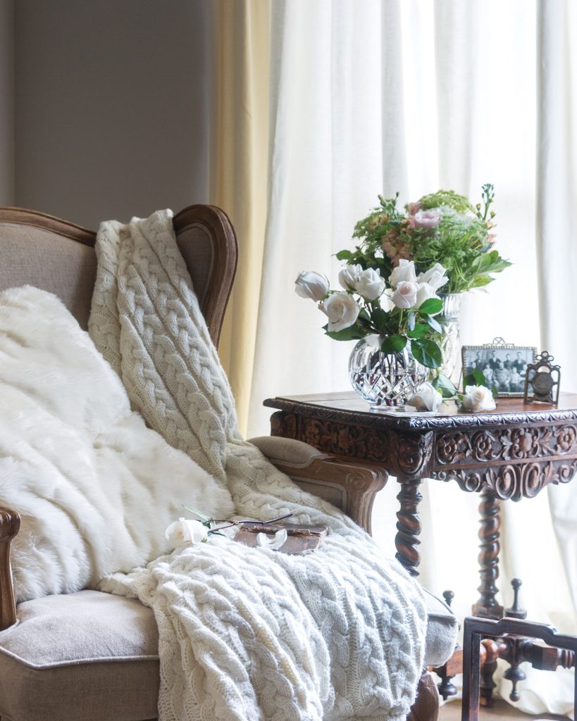 A soft reading nook sits beside the window, with a white chair dressed in cream quilt and pillow, beside a snowy display of flowers.