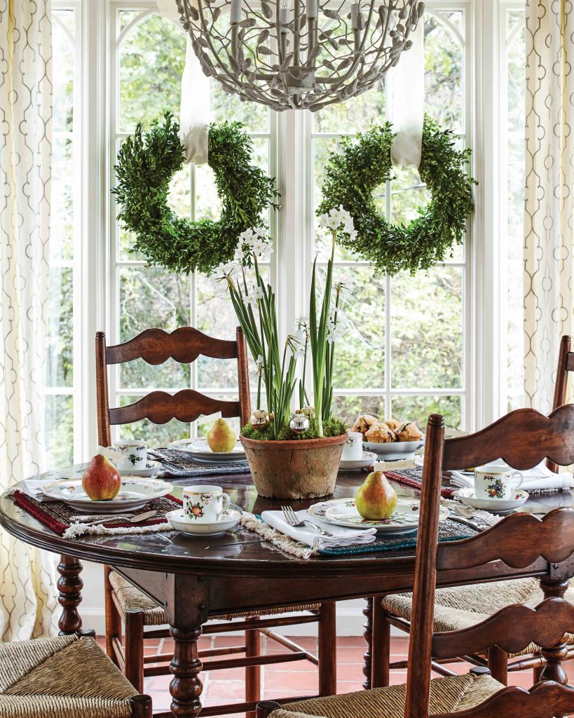 A breakfast table adorned with pears and paperwhites sits beside a window dressed in simple evergreen wreaths.