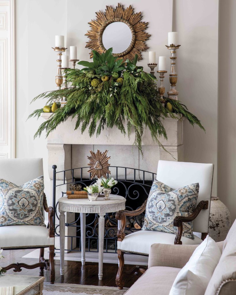 Branches of evergreen hang from atop an elegant hearth in a living room dressed in white and gold.