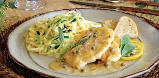 Served with a glass of white wine, zucchini noodles and chicken piccata are arranged on a plate beside slices of crunchy ciabatta.