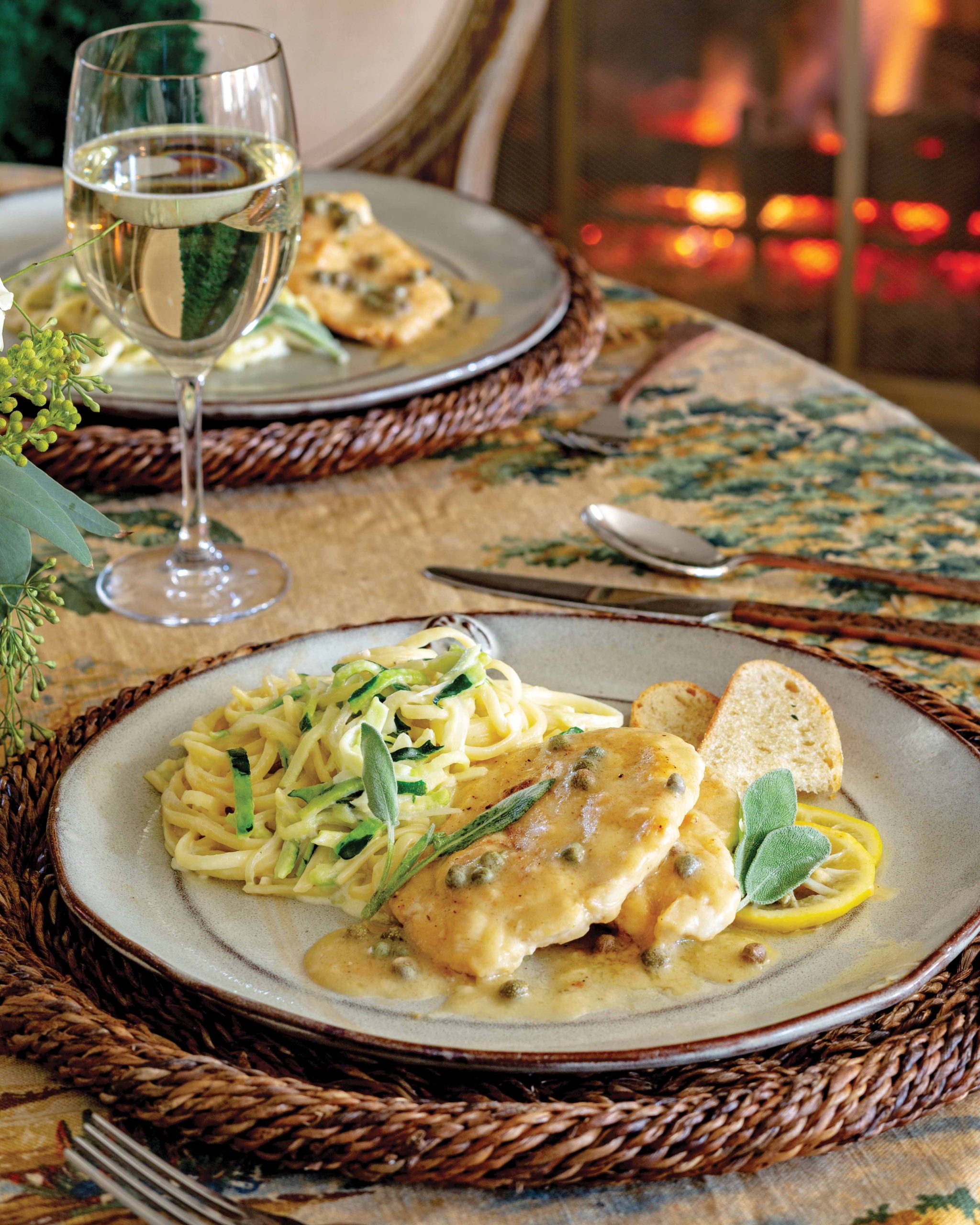 Served with a glass of white wine, zucchini noodles and chicken piccata are arranged on a plate beside slices of crunchy ciabatta.