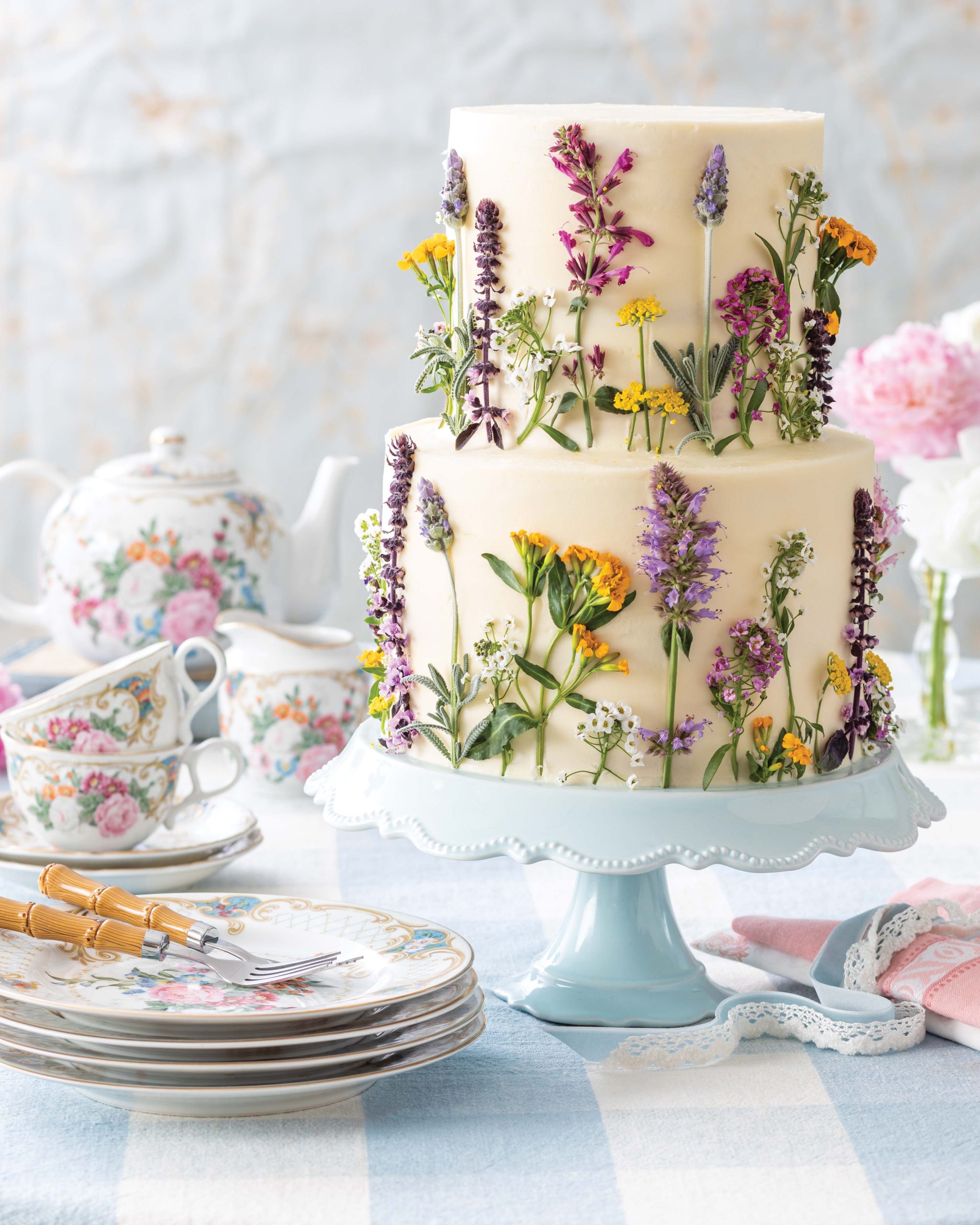 A blue and white checked table set for teatime with a white cake inlaid with edible blossoms atop a blue cake stand.