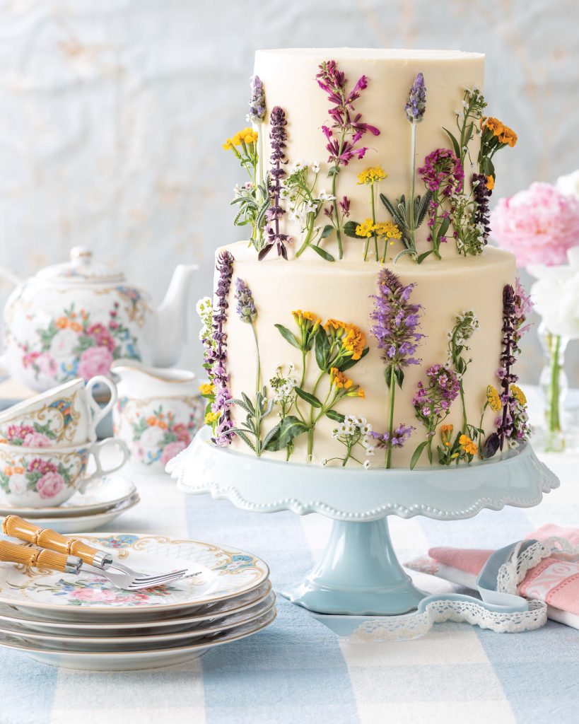 A two-tiered cake is frosted in white and adorned with real flowers in shades of purple and yellow.