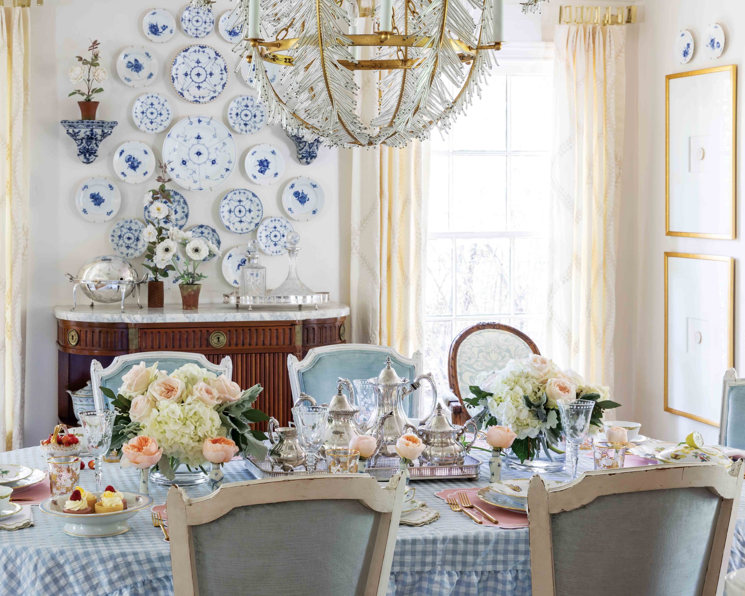 A blue-and-white room boasts a magnificent table, with pastel flowers, silver, and more awaiting a feast, while a collection of beautiful plates hangs on the wall.