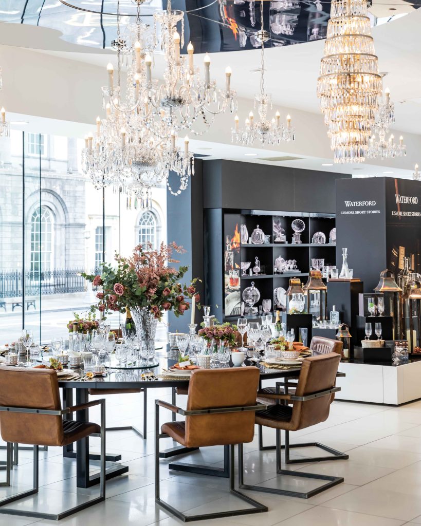 In the crystal maker’s storefront, magnificent chandeliers hang overhead while dining accoutrement are displayed atop a grand round table.