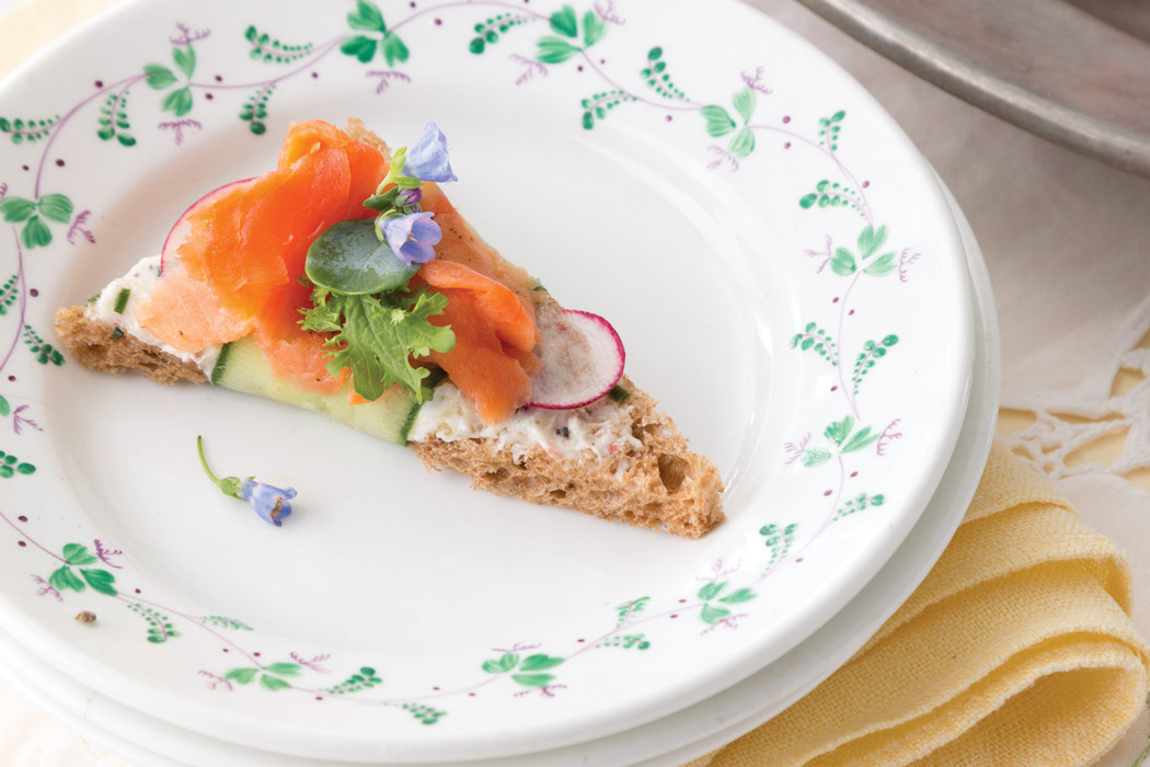 An open-faced Tea Sandwich with Edible Floral Compound Butter on a green leaf-rimmed white plate atop a yellow linen.