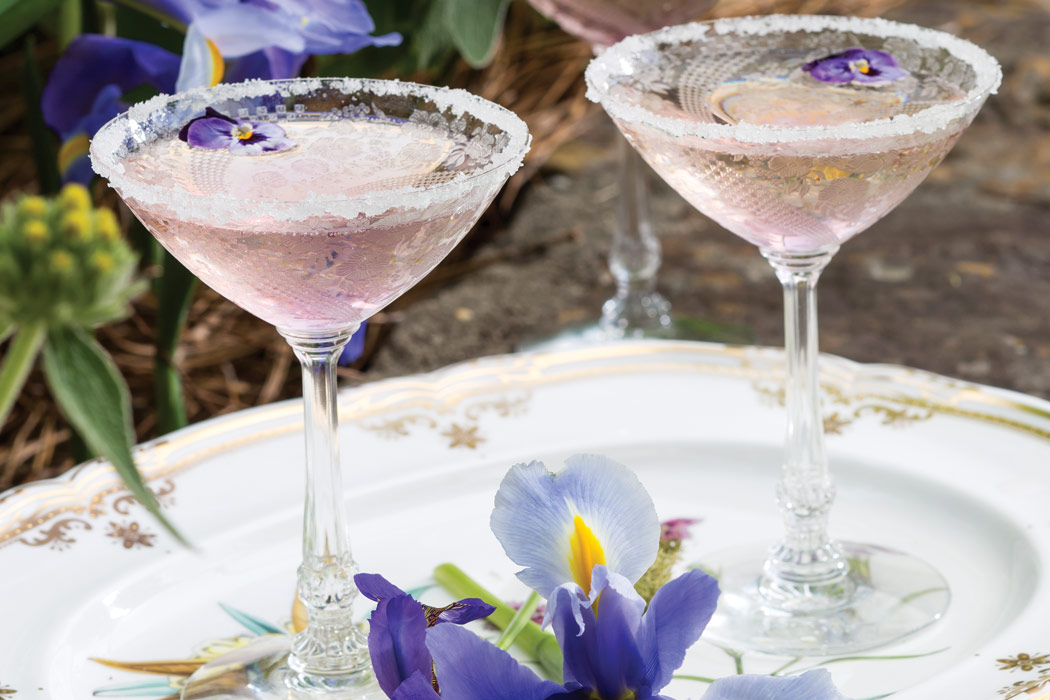A white and gold serving platter set with crystal goblets filled with violet champagne cocktail.