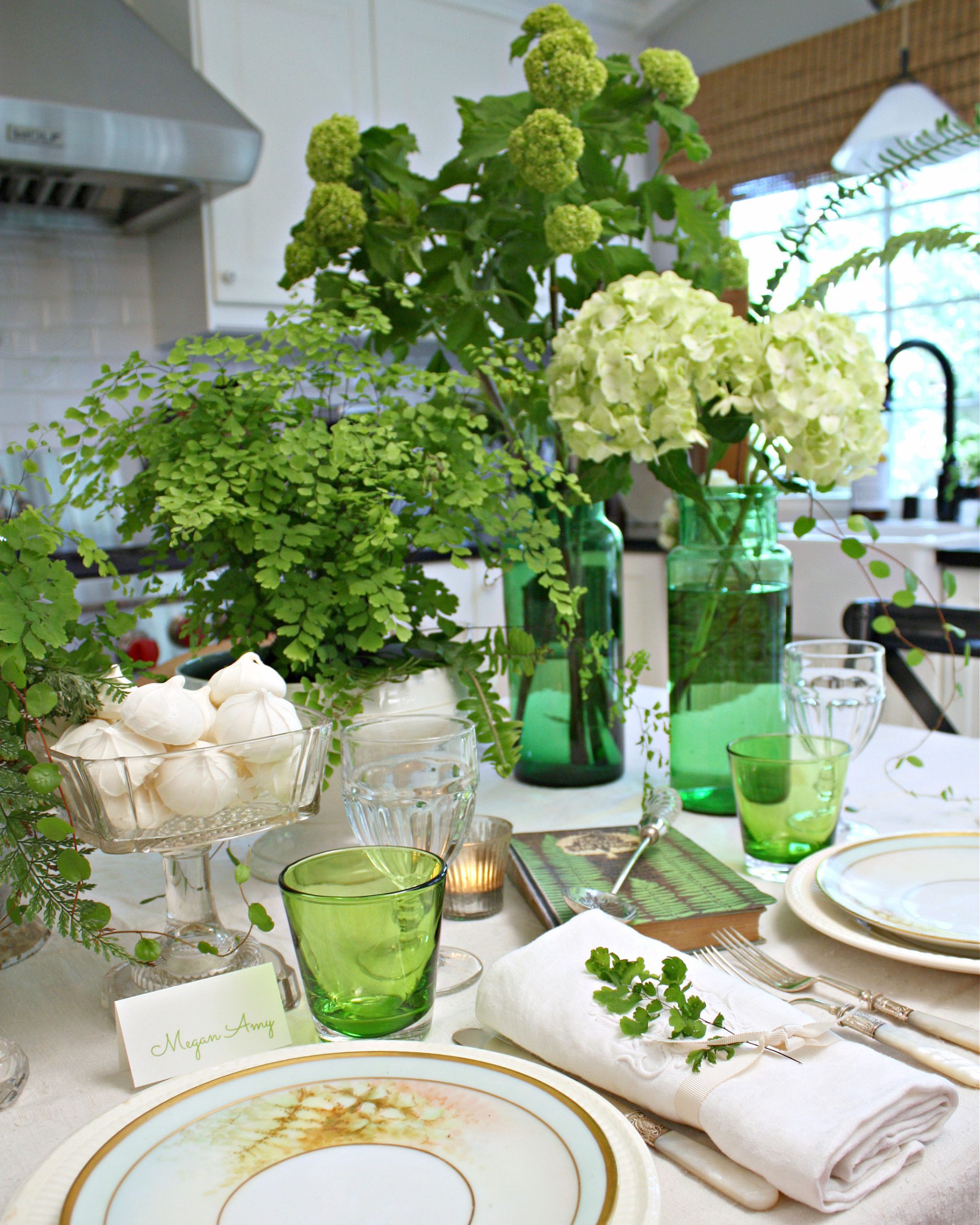 Set in the kitchen, this table has a collection of floral bouquets as its centerpiece, including green foliage and both green and white hydrangeas proudly displayed in tall green apothecary jars.