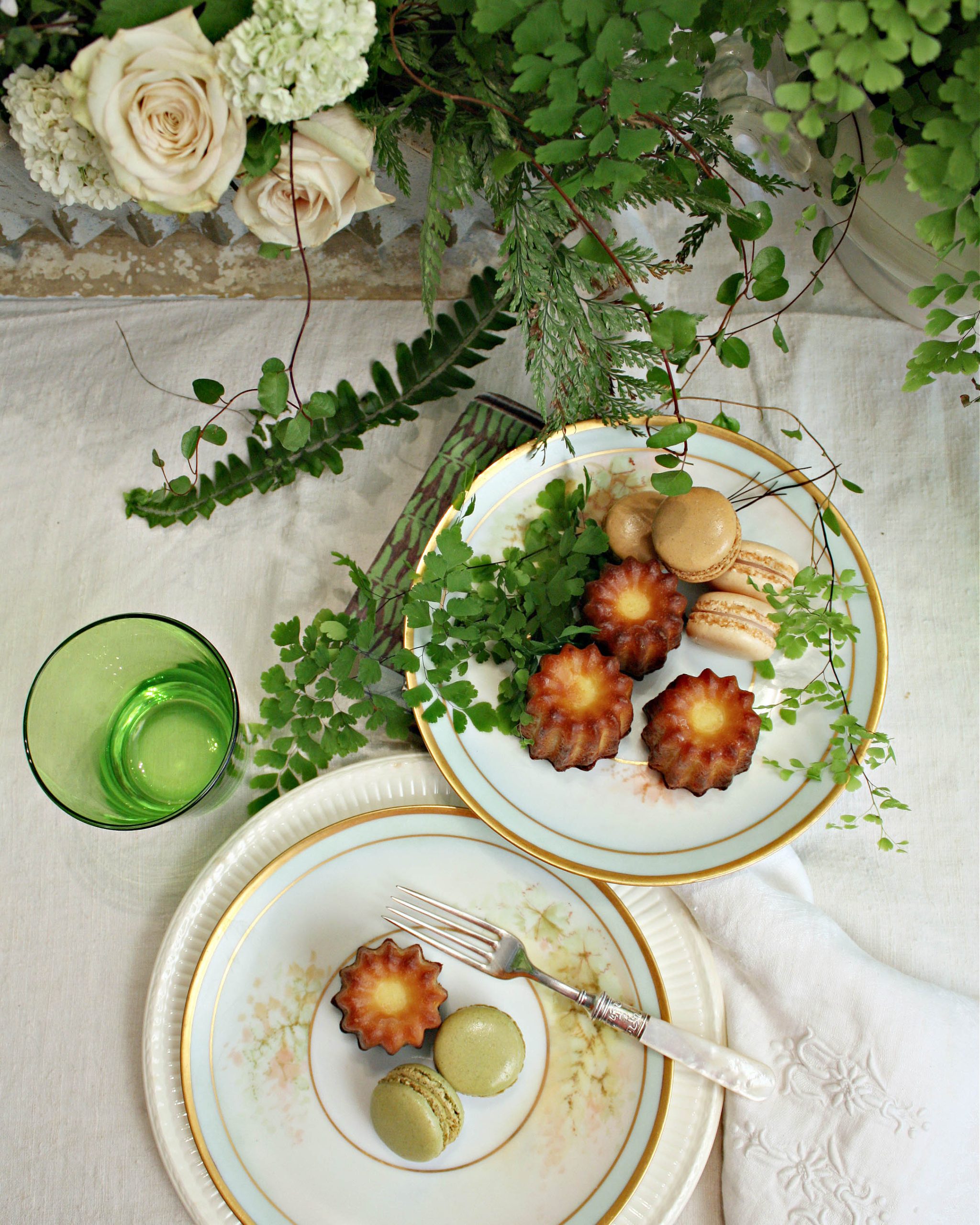 A collection of sweet pastries, such as macarons in shades of green and cream, rest atop plates painted with fronds of greenery.