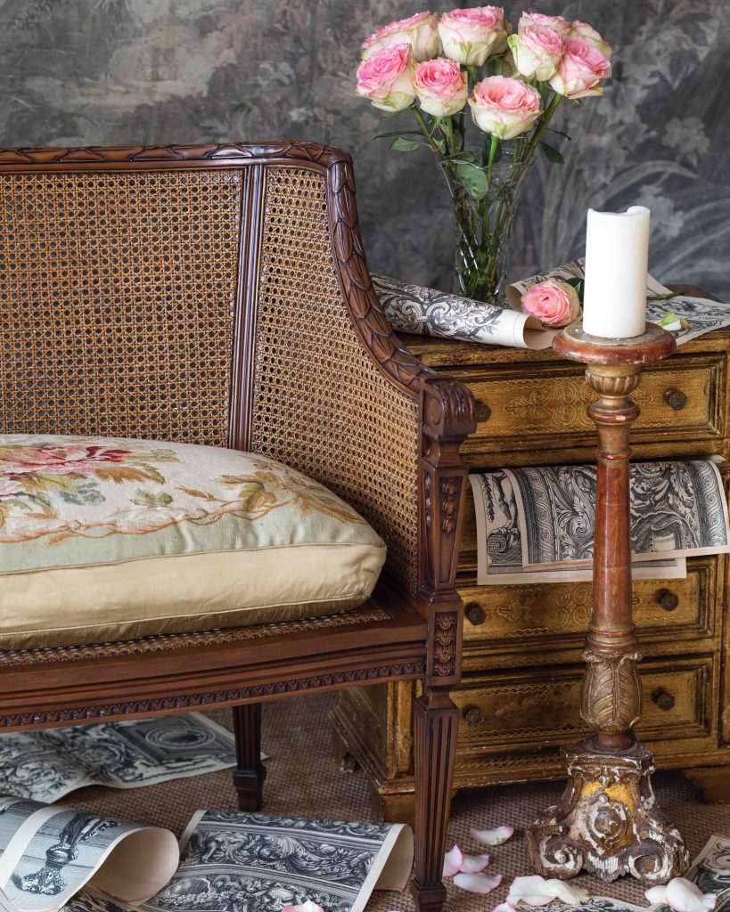 A wide cane seat with a floral-patterned cushion sits beside a dresser spilling with romantic French prints and rose petals.