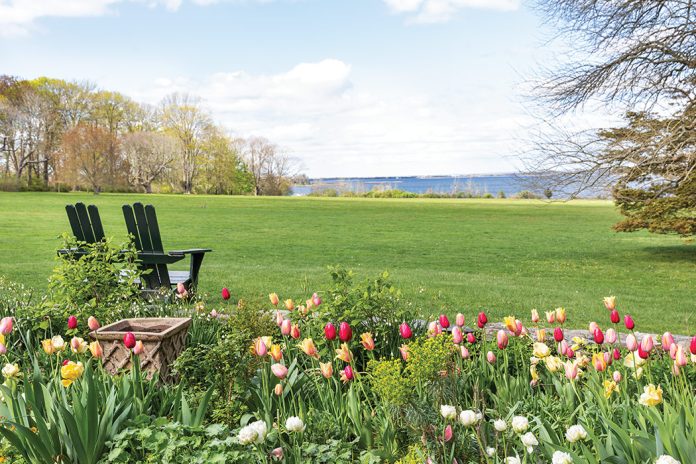 Two slender Adirondack chairs sit at the edge of a tulip garden and a verdant field, looking out onto the bay.