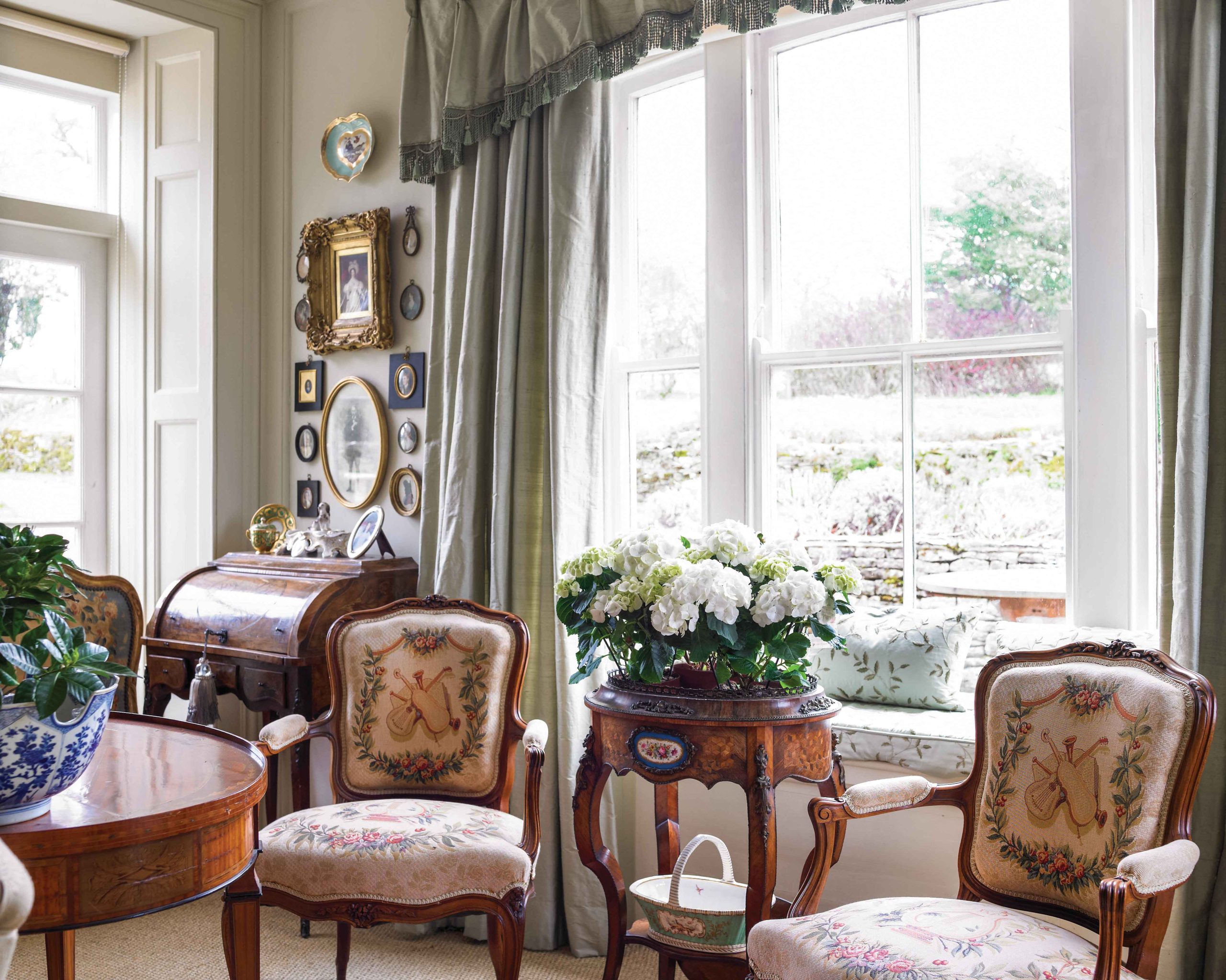 Beautiful needlework on the chair upholstery, small gilt frames upon the wall, and fresh hydrangeas all add elegance to this sitting area beside two bright windows.