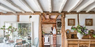 From Coast to Countryside: A breakfast nook with blue chairs sits beneath whitewashed wood ceilings and storied wooden furniture, with a peek through the open doorway into the cozy kitchen.