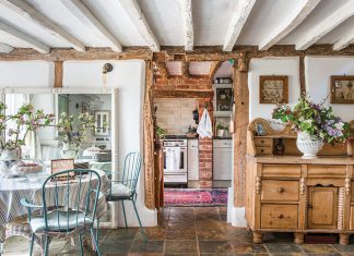 From Coast to Countryside: A breakfast nook with blue chairs sits beneath whitewashed wood ceilings and storied wooden furniture, with a peek through the open doorway into the cozy kitchen.