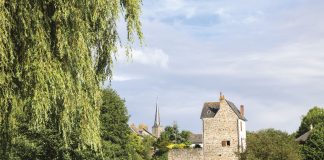 A calming view of the French countryside, with a village of stone buildings set beside a river bank and beneath a brilliant blue sky.