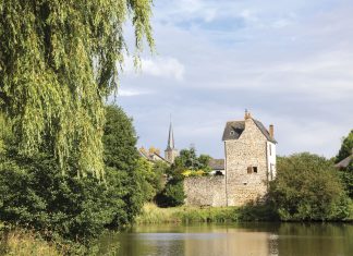 A calming view of the French countryside, with a village of stone buildings set beside a river bank and beneath a brilliant blue sky.
