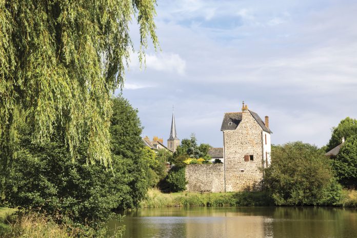 A calming view of the French countryside, with a village of stone buildings set beside a river bank and beneath a brilliant blue sky.