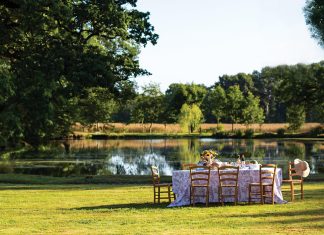 In a grassy clearing beside a sparkling lake, a breezy table is set for summer dining