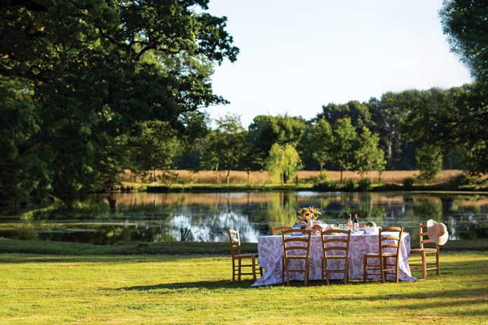 In a grassy clearing beside a sparkling lake, a breezy table is set for summer dining