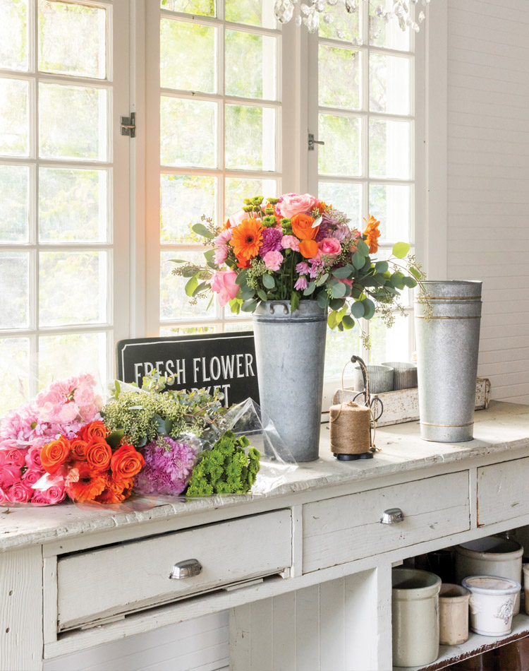 Leslie Saeta has a designated nook for arranging flowers that she refreshes around the home each week. Colorful blooms of orange, pink, and purple are stacked on the potting bench ready to be placed in a tall metal can.