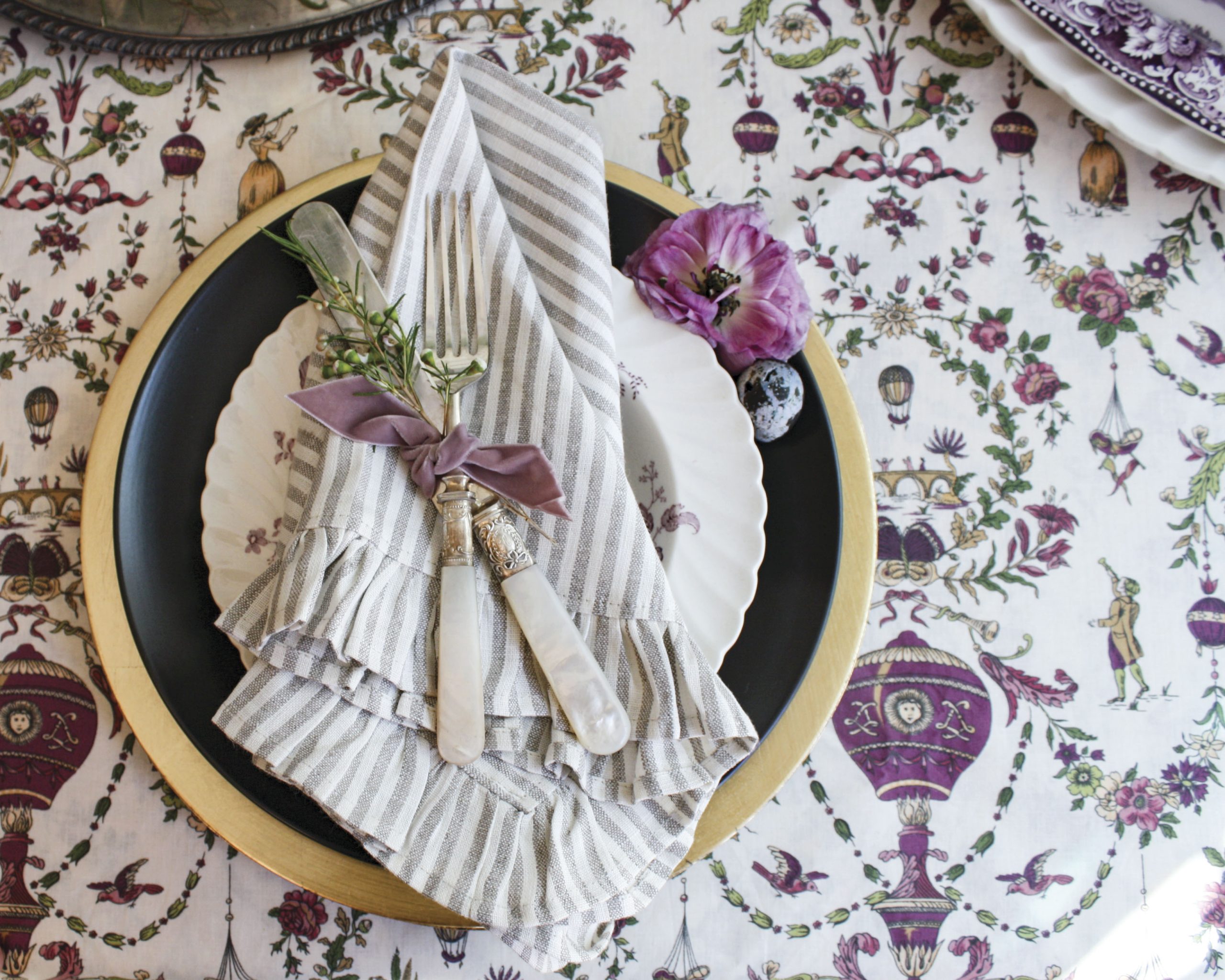 An ornate white-and-purple tablecloth makes quite a statement and is dressed in a mix of modern and antique items in this place setting, including a gold charger, black plate, and striped linen napkin.