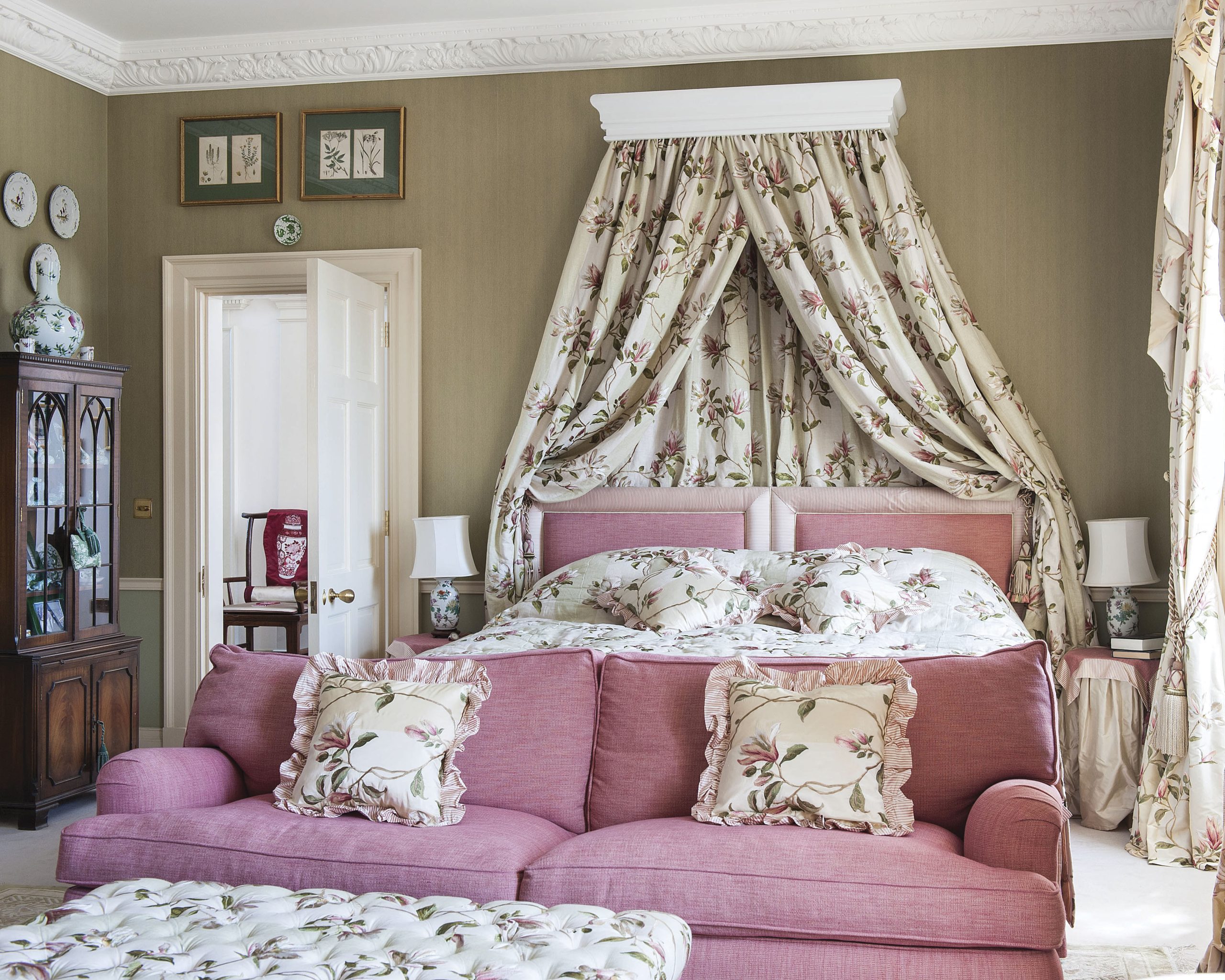 A pink sofa and headboard pair nicely with the floral fabrics of this bedroom, which boasts a storied feminine atmosphere.