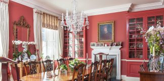 Red walls and cupboards bring a bold atmosphere to this grand dining room, which seats ten people amid framed portraits, cherished antiques, and fine textiles.