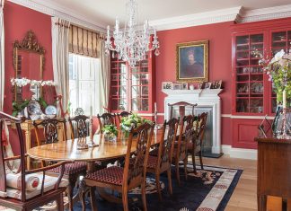 Red walls and cupboards bring a bold atmosphere to this grand dining room, which seats ten people amid framed portraits, cherished antiques, and fine textiles.