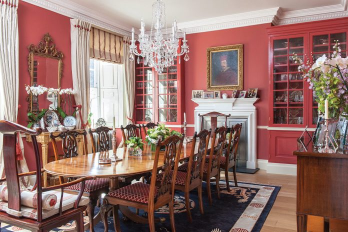 Red walls and cupboards bring a bold atmosphere to this grand dining room, which seats ten people amid framed portraits, cherished antiques, and fine textiles.